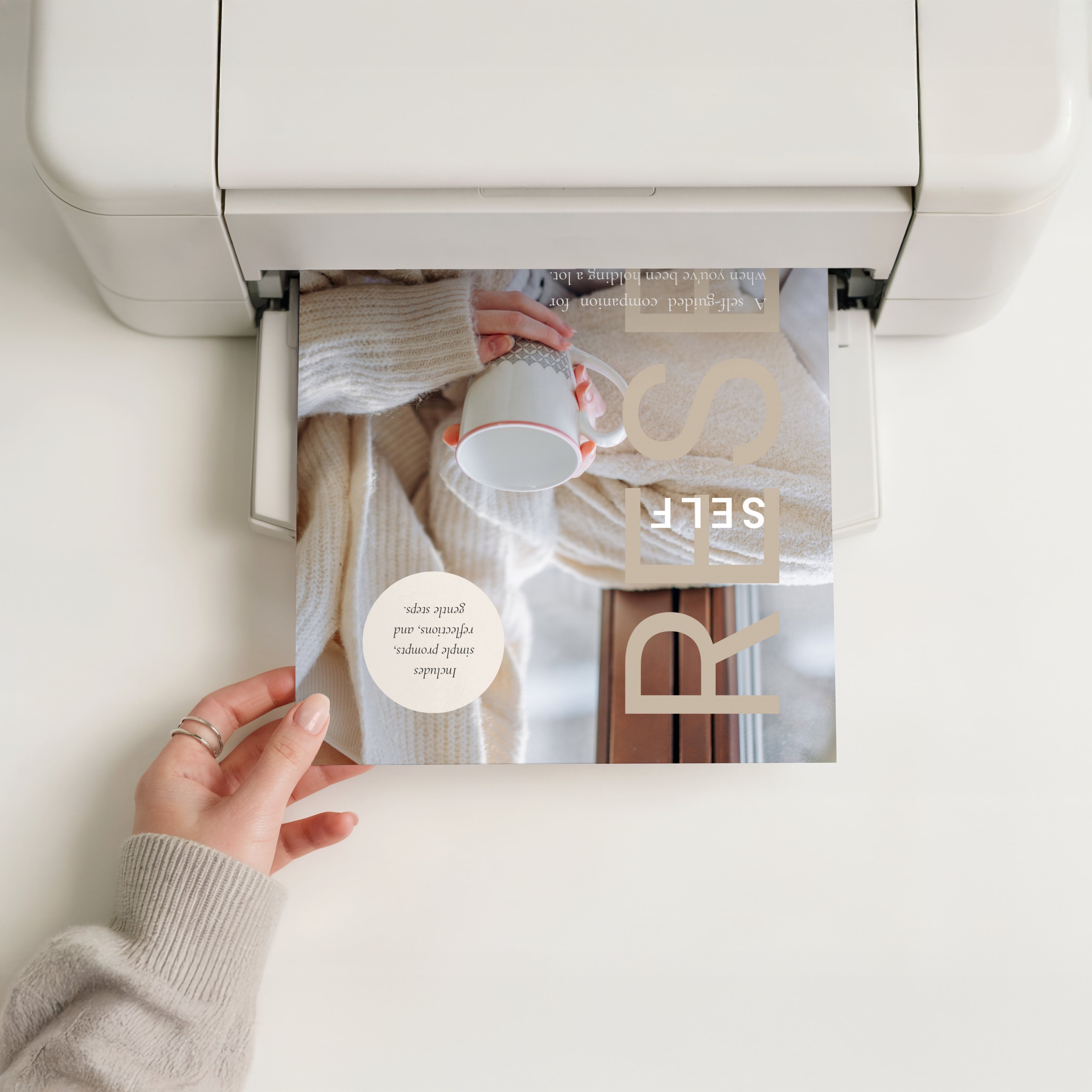 Hand reaching out and holding the cover of The Self Reset as it comes out of a personal printer at home